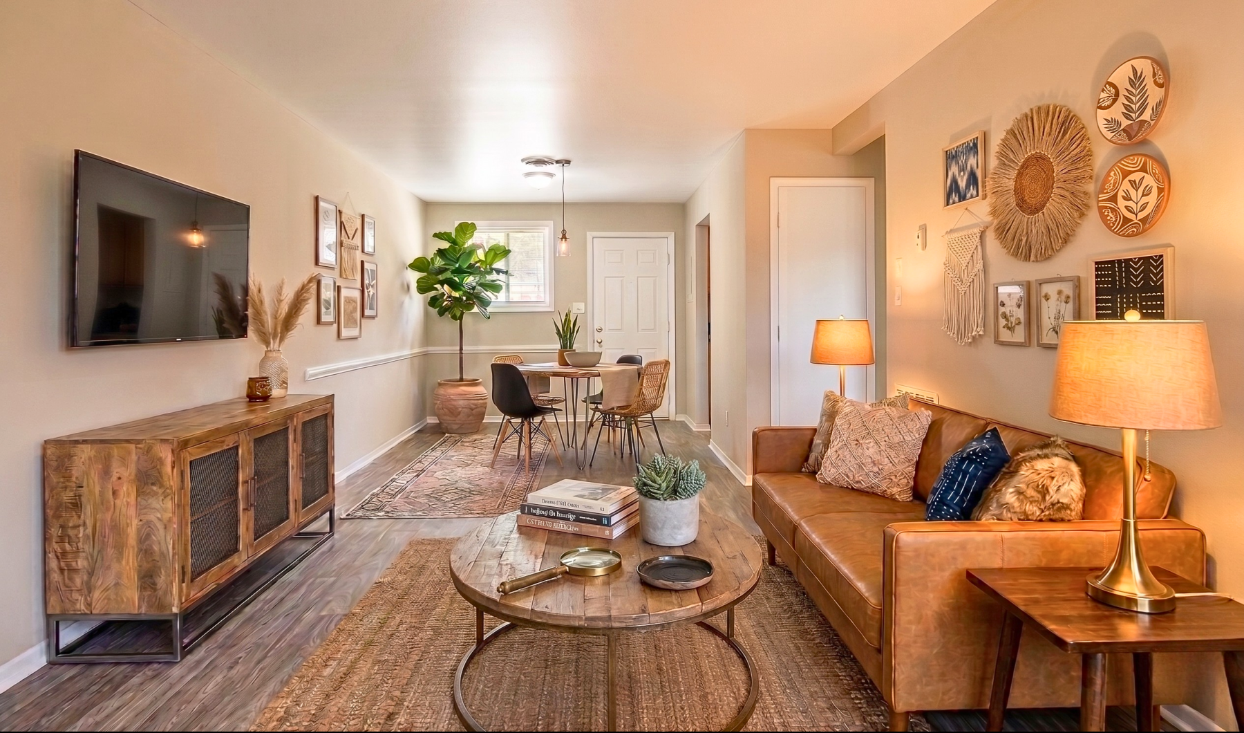 Warmly decorated apartment living room with a cognac leather sofa, round wood coffee table, wall-mounted TV, and an open dining area.